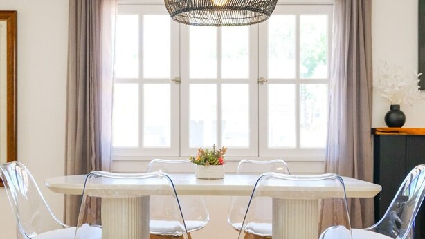 A dining area with a round table, four clear chairs, a woven light fixture, and a window with curtains. - Silver Palms - Holiday in Style: Beach + Pool! (St. Augustine Beach)