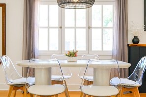A dining area with a round table, four clear chairs, a woven light fixture, and a window with curtains. - Silver Palms - Holiday in Style: Beach + Pool! (St. Augustine Beach)