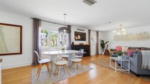 A spacious living room with a grey sofa, a dining table with chairs, a TV mounted on the wall, and a large window with curtains. - Silver Palms - Holiday in Style: Beach + Pool! (St. Augustine Beach)