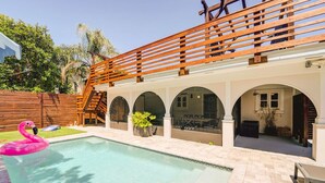 A swimming pool with a wooden deck and arched openings, surrounded by a wooden fence and greenery. - Silver Palms - Holiday in Style: Beach + Pool! (St. Augustine Beach)