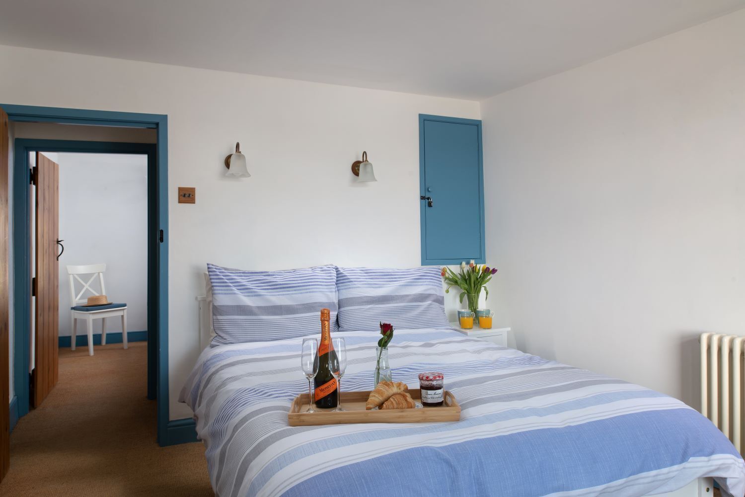 A bedroom with a bed, a chair, a tray with a bottle, glasses, a rose, and a small table with a vase and orange juice.