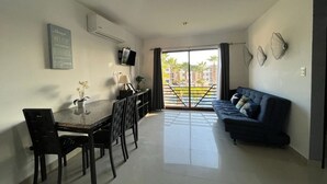 Uma sala de estar moderna com um sofá azul escuro, uma mesa de jantar com cadeiras e uma TV fixada na parede. É visível uma varanda com vista para palmeiras e edifícios através das portas de vidro.