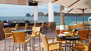 An outdoor seating area with wicker chairs and tables, overlooking a body of water with rocky formations in the background.