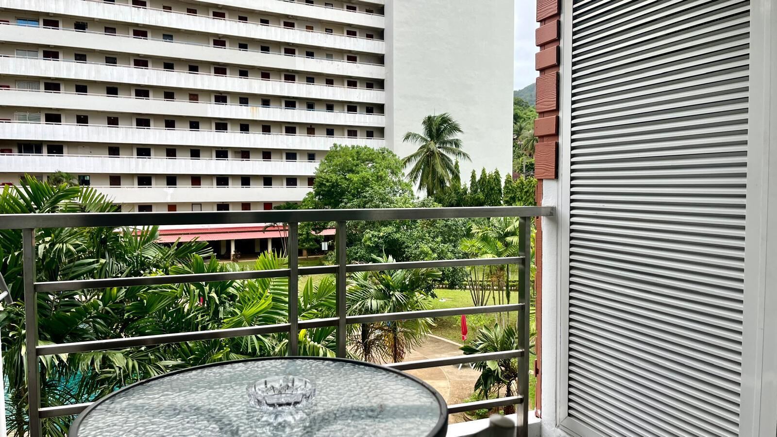 A balcony with a glass table, surrounded by lush greenery and a view of a multi-story building.