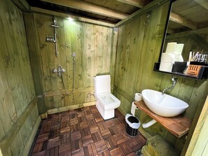 A rustic wooden bathroom with a wooden floor, a white toilet, a shower with a handheld showerhead, and a sink with a mirror above it. - Ala Glamping (Mudurnu)