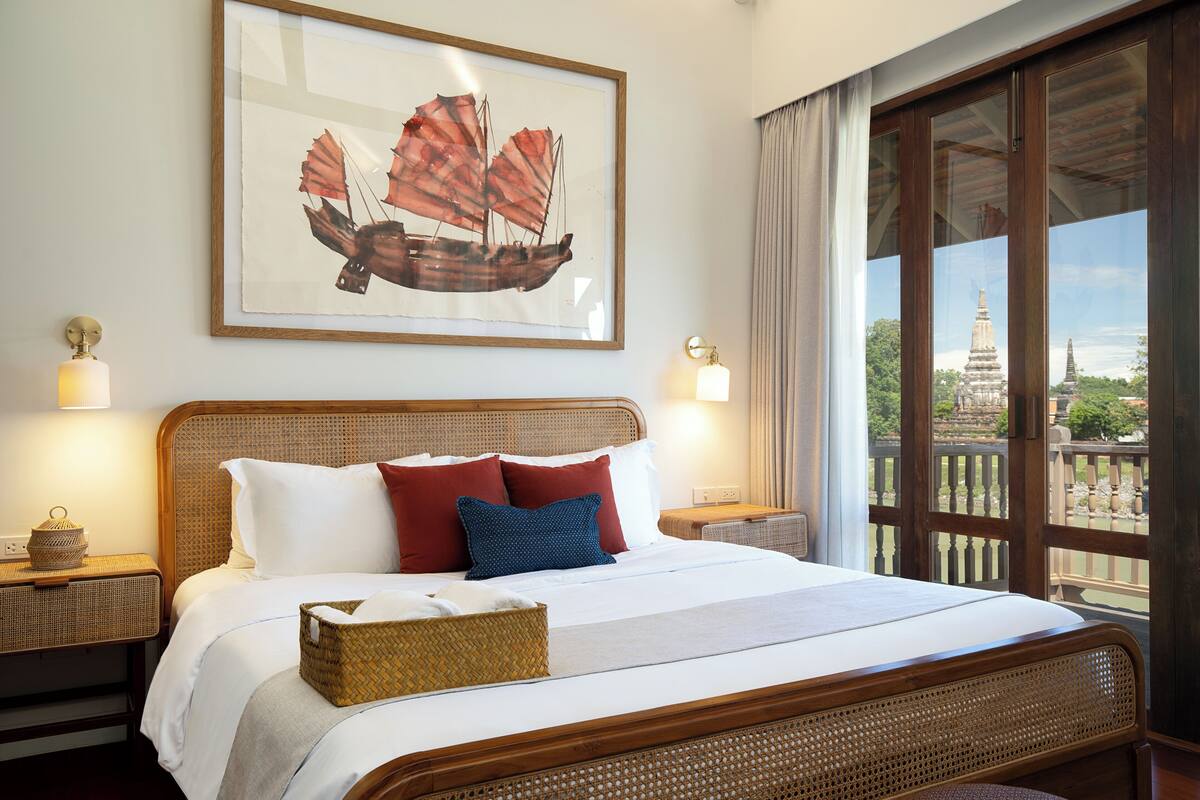 Habitación de hotel con cama, un cuadro de un barco, dos lámparas de noche y vista a un templo a través de la ventana.
