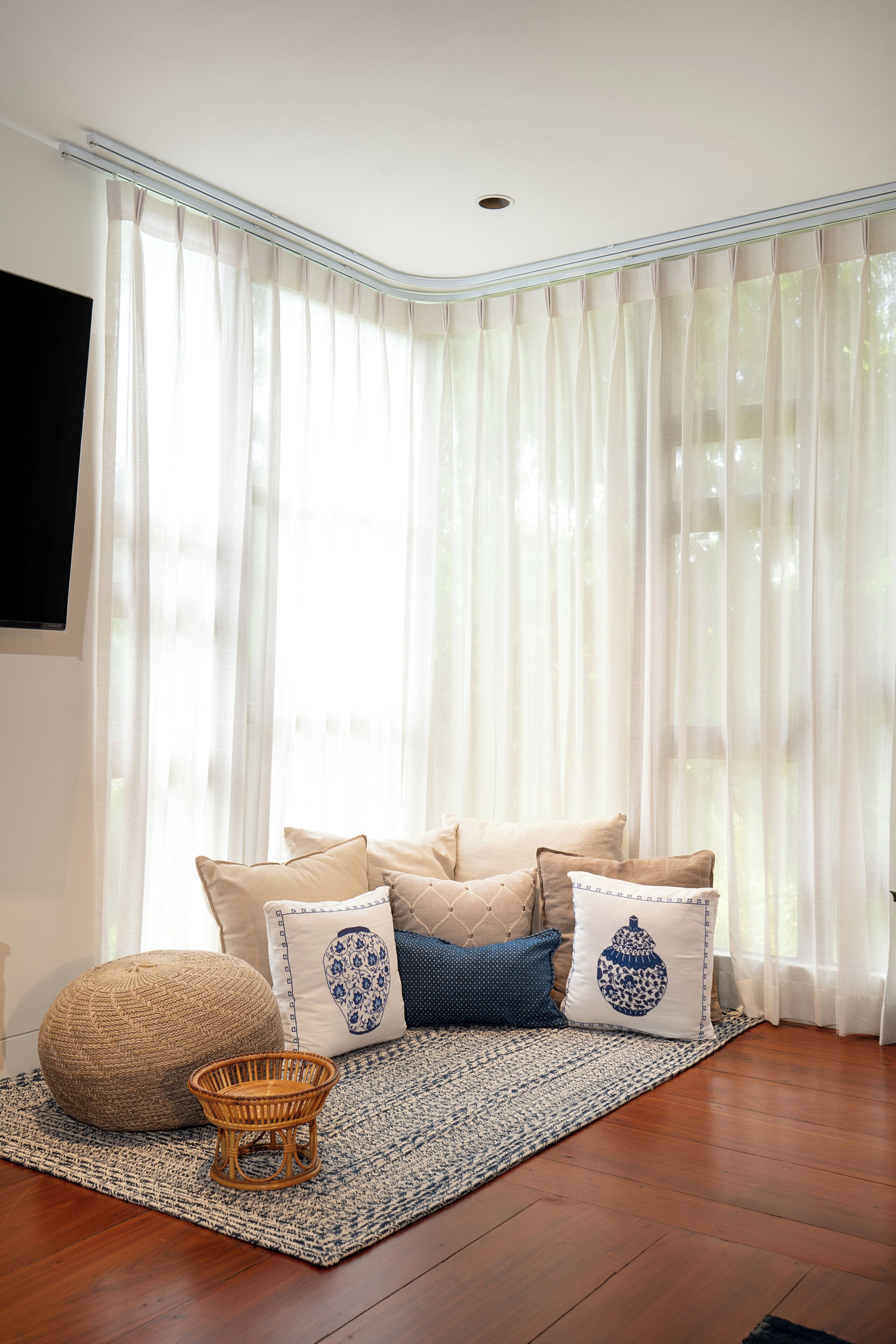 Une pièce d’angle avec une grande fenêtre, un canapé agrémenté de coussins à motifs, un pouf en fibres tressées et une petite chaise en osier.