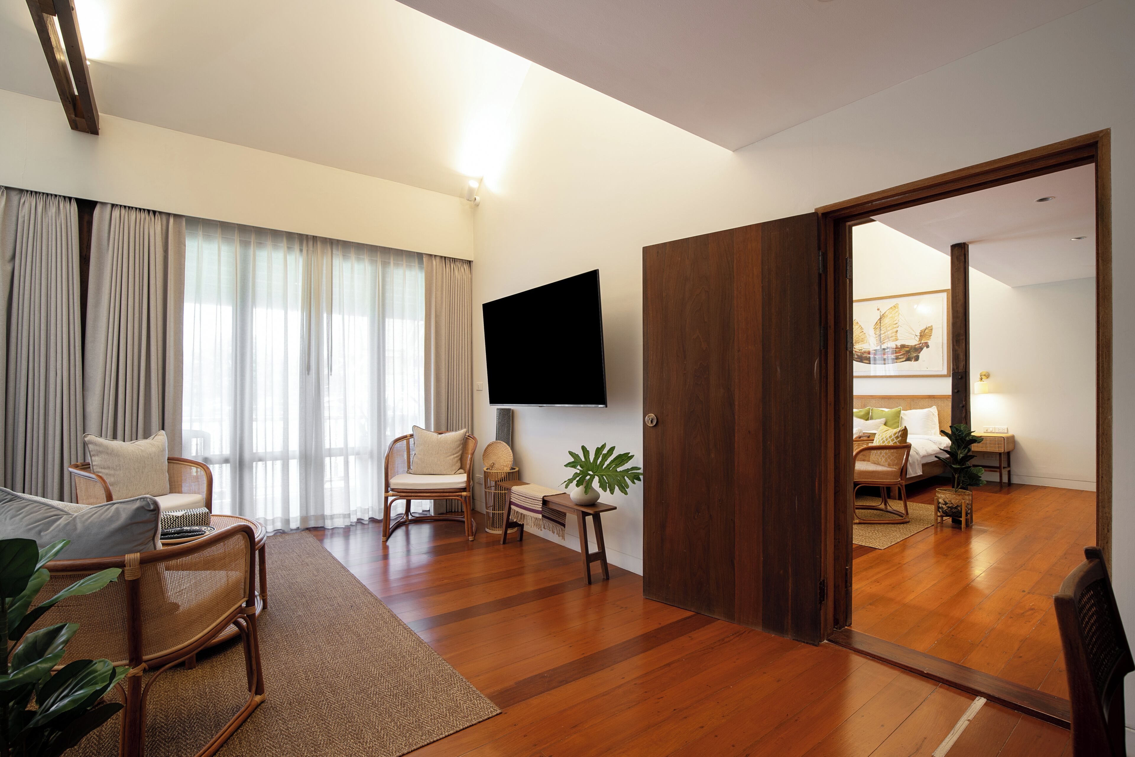 A modern living room with a wooden floor, a flat-screen TV mounted on the wall, and a sliding door leading to another room.