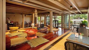 A spacious indoor area with wooden flooring, a central seating area with a table and chairs, and large windows offering a view of the outdoors. - Syama Residence (Ayutthaya)