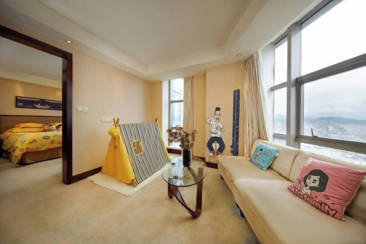 A hotel room with a large window, a sofa with a cartoon pillow, a glass coffee table, a bed with a yellow blanket, and a wall painting.
