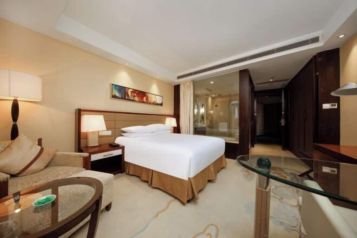 A hotel room with a bed, a sofa, a glass table, a nightstand, and a painting on the wall.