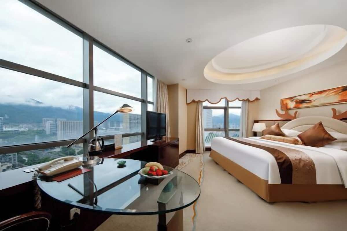 A hotel room with a large window, a glass table, a bed with pillows, and a painting on the wall.