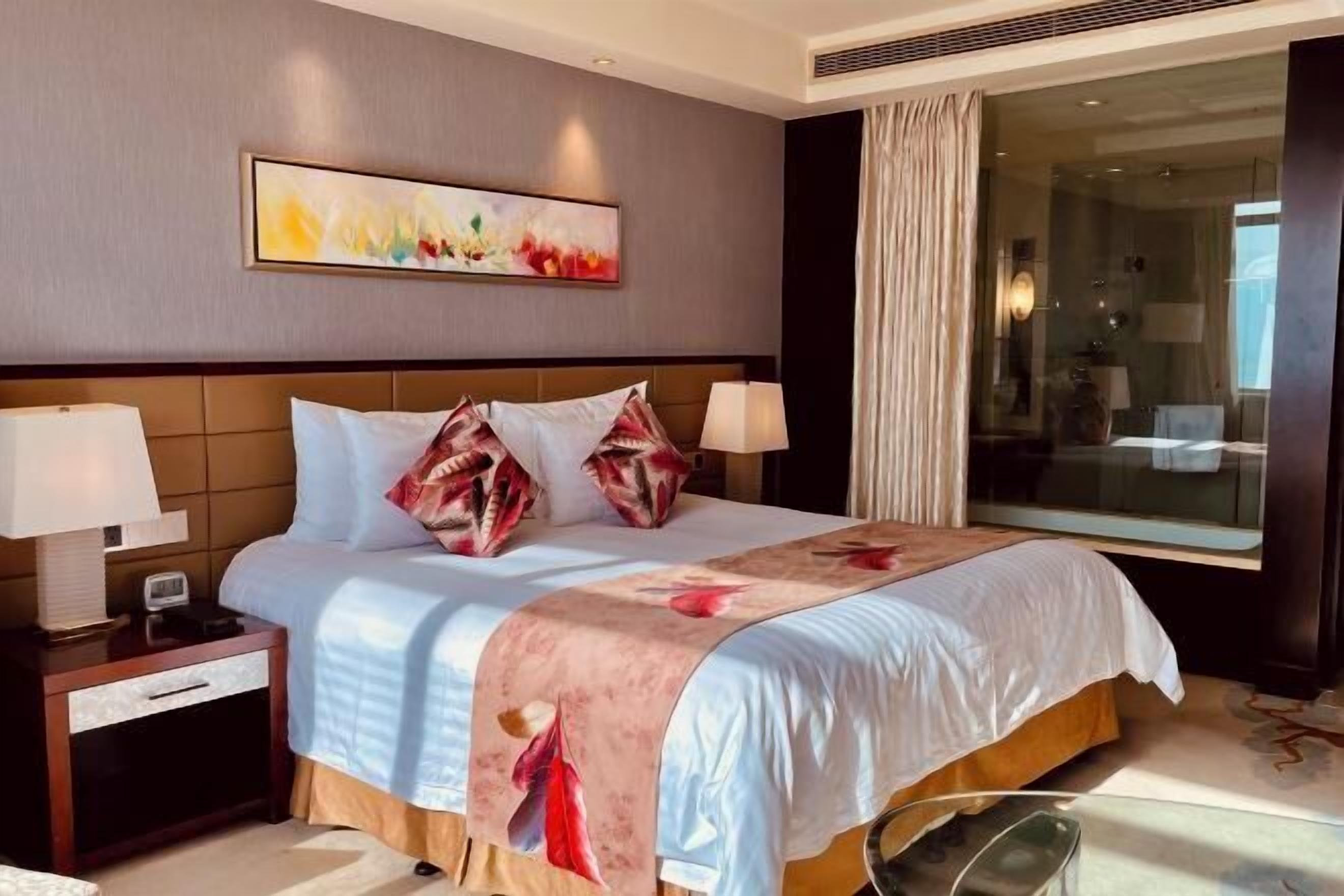 A hotel room with a large bed, two bedside lamps, a nightstand, a glass shower enclosure, and a large window with curtains.