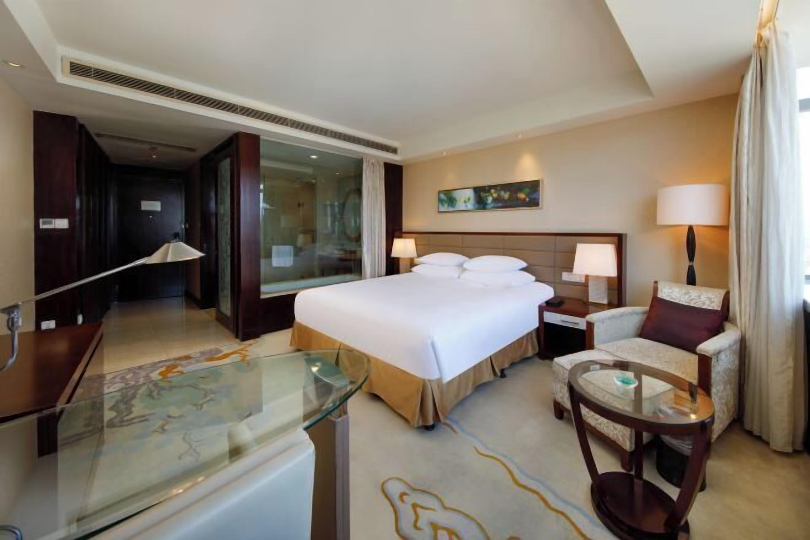 A hotel room with a large bed, a glass desk, a chair, a nightstand, and a painting on the wall.