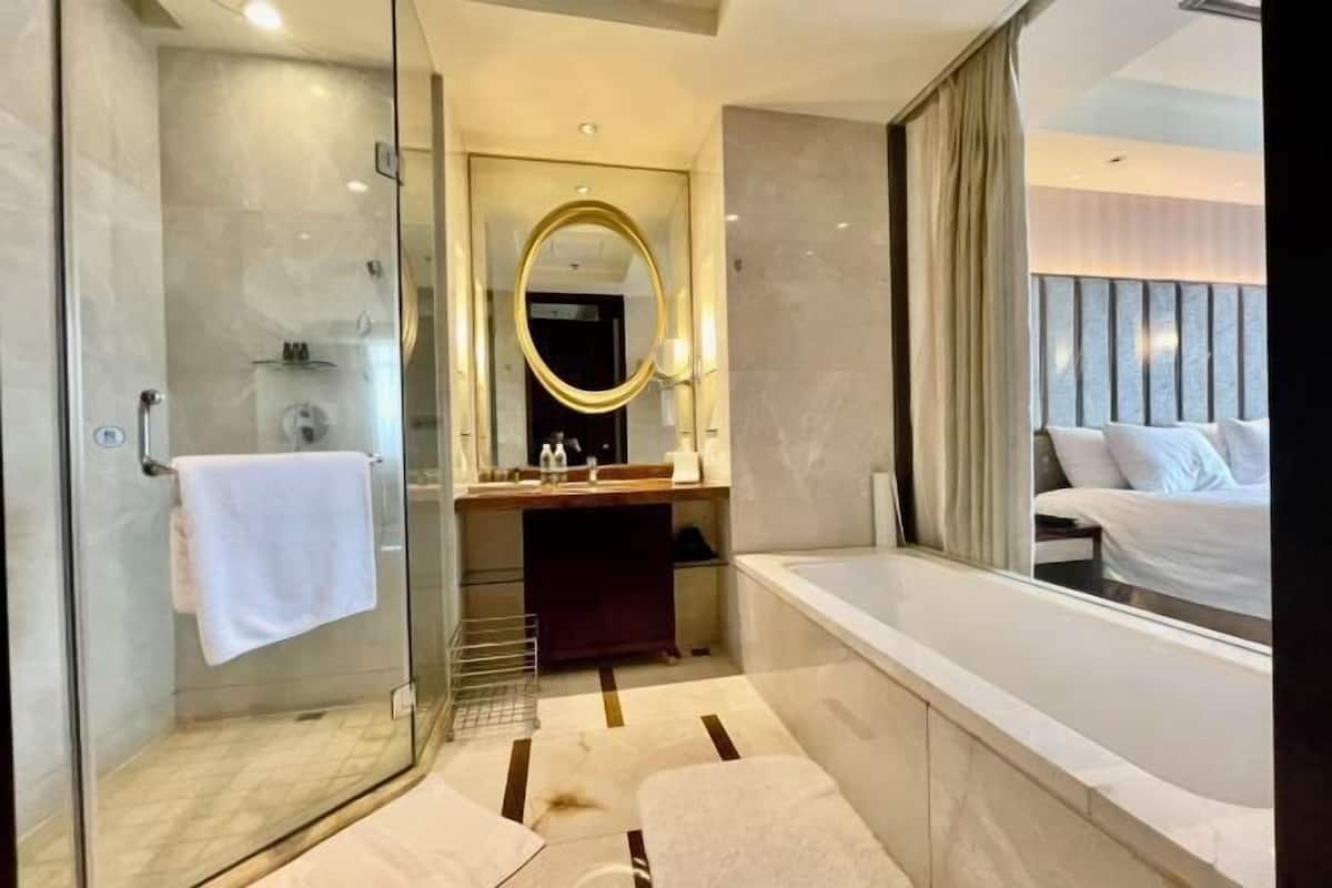A modern bathroom with a glass shower, a large mirror, a bathtub, and a view of a bedroom with a bed.