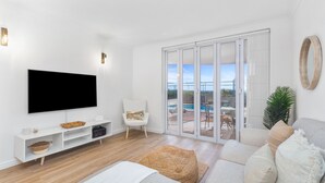 A modern living room with a flat-screen TV, a white armchair, a sofa with cushions, a wooden floor, and a sliding glass door leading to an outdoor area.