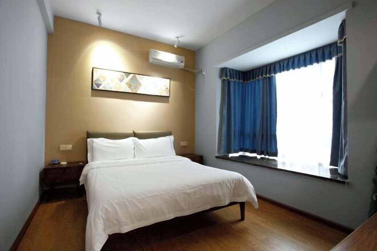 A bedroom with a bed, a nightstand, an air conditioning unit, a window with curtains, and a wall-mounted picture.