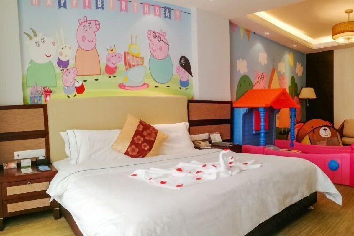 A hotel room with a bed, pillows, a nightstand, a lamp, a wall mural, and a play area.