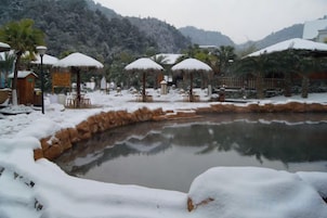 A snow-covered outdoor area with a pond, thatched-roof structures, and a mountainous backdrop.