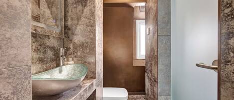 A bathroom with a glass vessel sink, a white toilet, and a marble wall.
