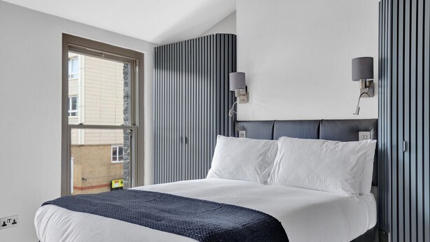 Une chambre d’hôtel moderne avec un grand lit, une fenêtre donnant sur des immeubles, une lampe fixée au mur et un panneau décoratif à rayures.