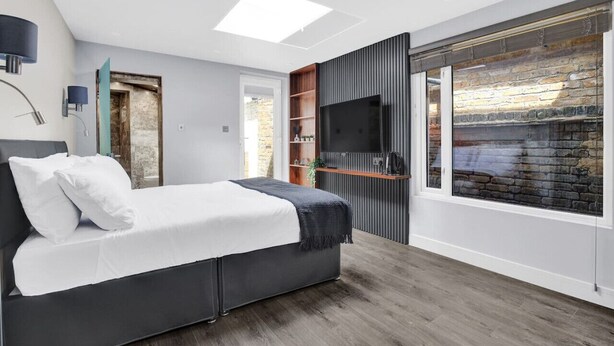 Une chambre à coucher moderne avec un lit, une télévision, une étagère en bois et une grande fenêtre donnant sur un mur de briques.