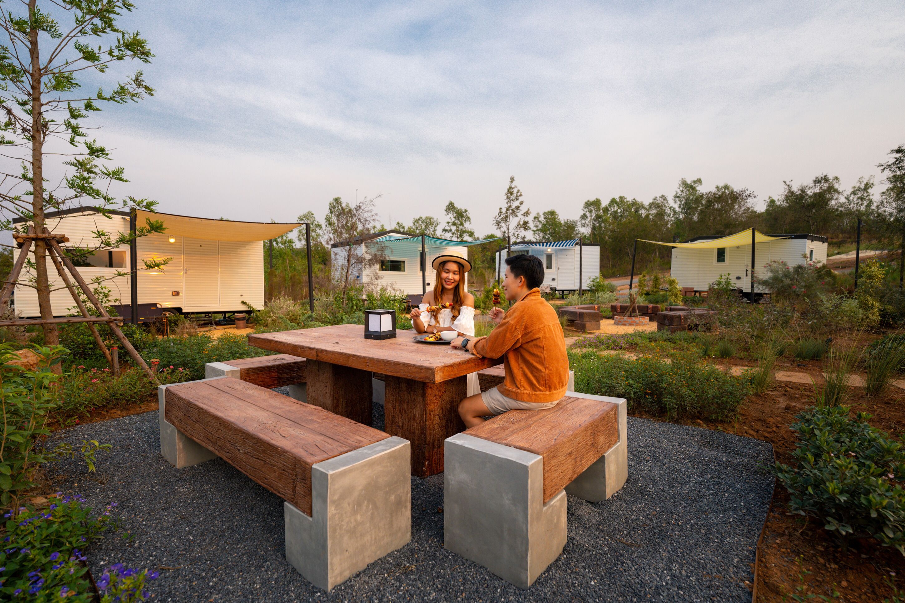 A wooden picnic table with benches, surrounded by gravel and greenery, with mobile homes in the background.