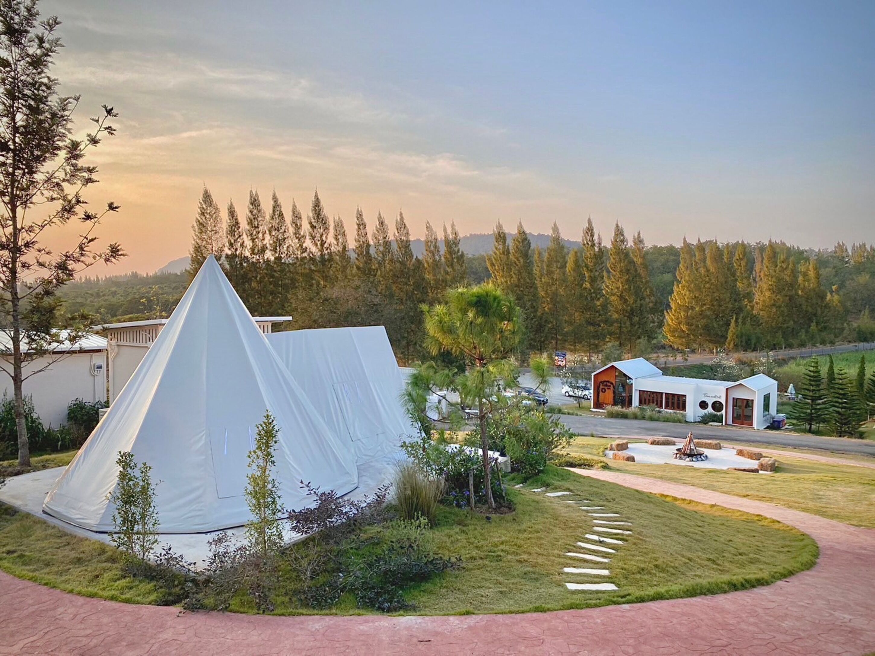 A tented accommodation with a paved driveway, a small building with a red door, and a view of a forested area.