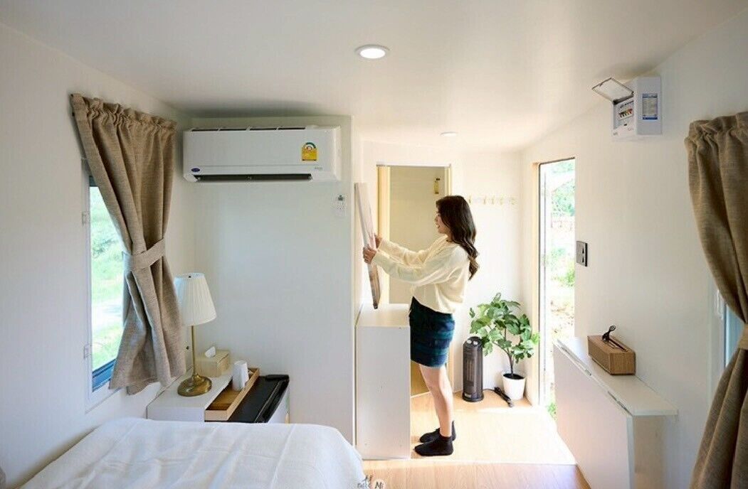A hotel room with a bed, a desk with a lamp, a potted plant, and an air conditioning unit.
