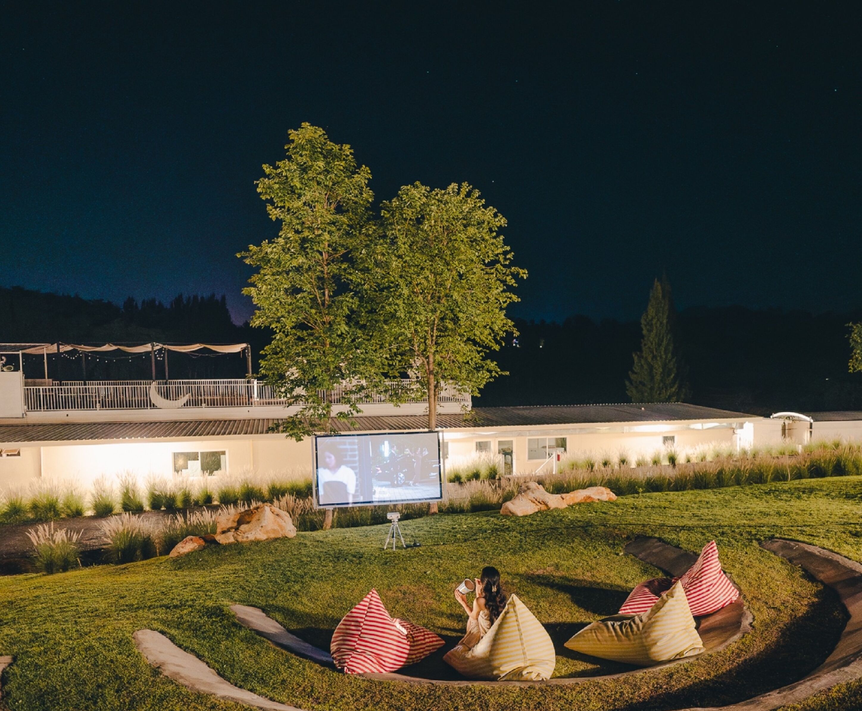 A nighttime outdoor seating area with a large screen, trees, and a building in the background.