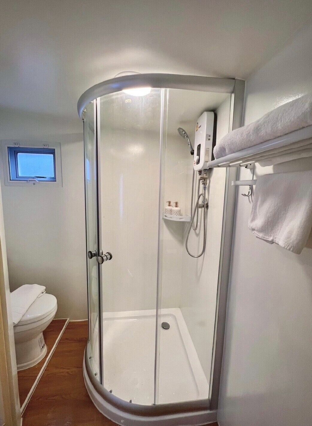 A glass-enclosed shower with a handheld showerhead, a white toilet, and a small window.