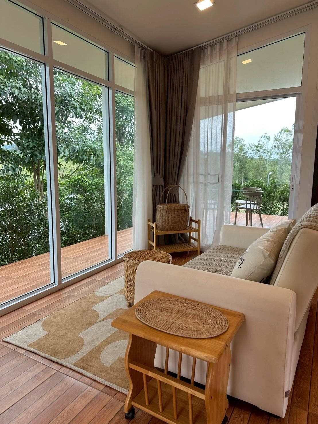 A modern living room with a sofa, a small wooden table, and a wicker chair. Large windows offer a view of greenery outside.