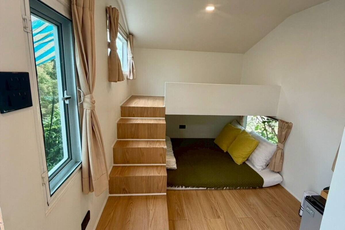 Une petite pièce avec un lit superposé, un escalier en bois, une fenêtre avec des rideaux et un luminaire au plafond.