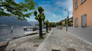 A stone-paved waterfront walkway with boats docked, trees, and a building with shutters. - Piazza Campidoglio 5 (Tremezzina)