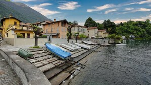 A lakeside village with boats docked along a stone embankment, surrounded by traditional buildings and lush greenery.