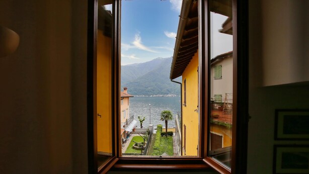 A view through an open window of a courtyard with a mountain in the background, a body of water, and buildings with balconies.