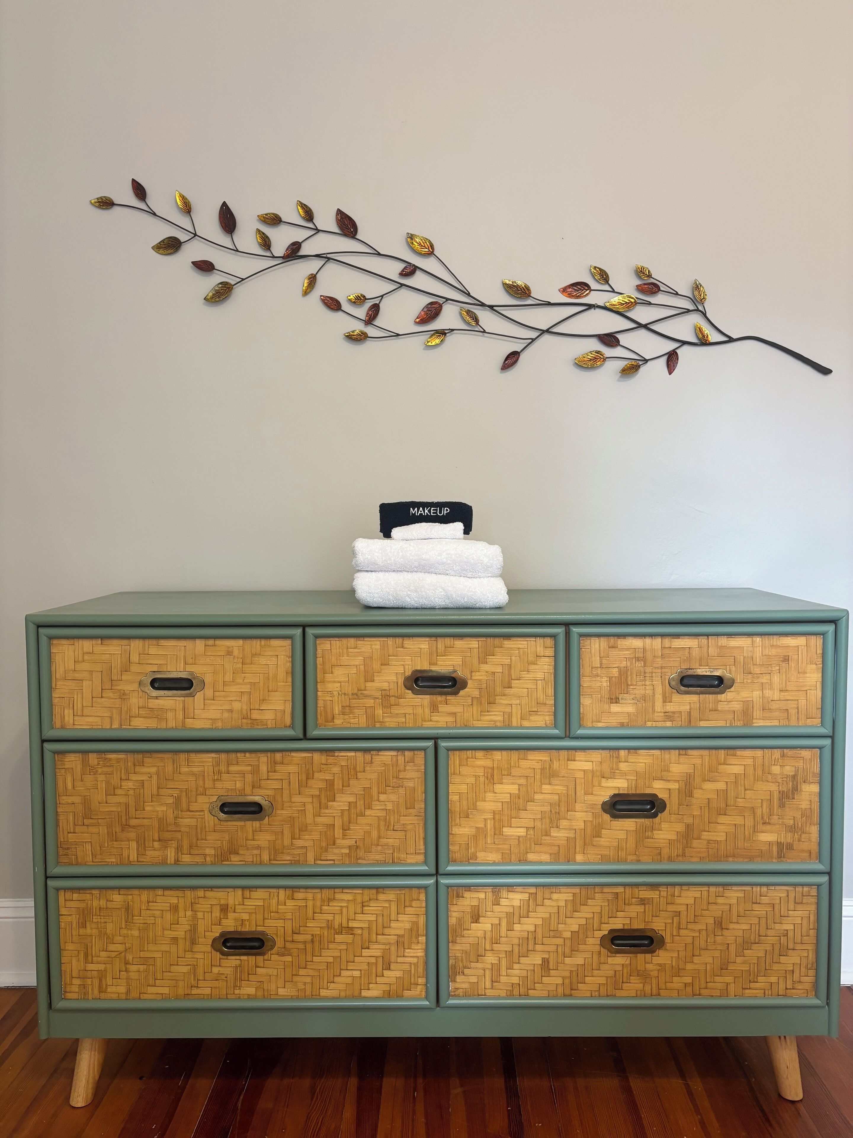 A teal-colored chest of drawers with a woven bamboo pattern, featuring six drawers and two doors, each with a metal handle. A decorative branch with leaves is mounted on the wall above the chest.