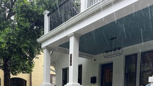 A white building with a covered porch, steps leading to a door, and a garden area with plants and flowers.