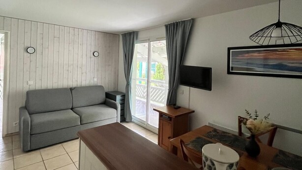 A modern living room with a grey sofa, a wooden dining table, and a kitchen area with a sink and cabinets.