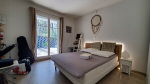 Ein Schlafzimmer mit einem großen Bett, einem Schreibtisch, einer Gitarre und Blick durch eine Schiebetür auf einen Außenbereich.
