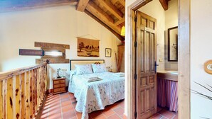 Un dormitorio con cabecera de madera, cama con edredón floral, mesita de noche con lámpara y puerta de madera que da a un baño.