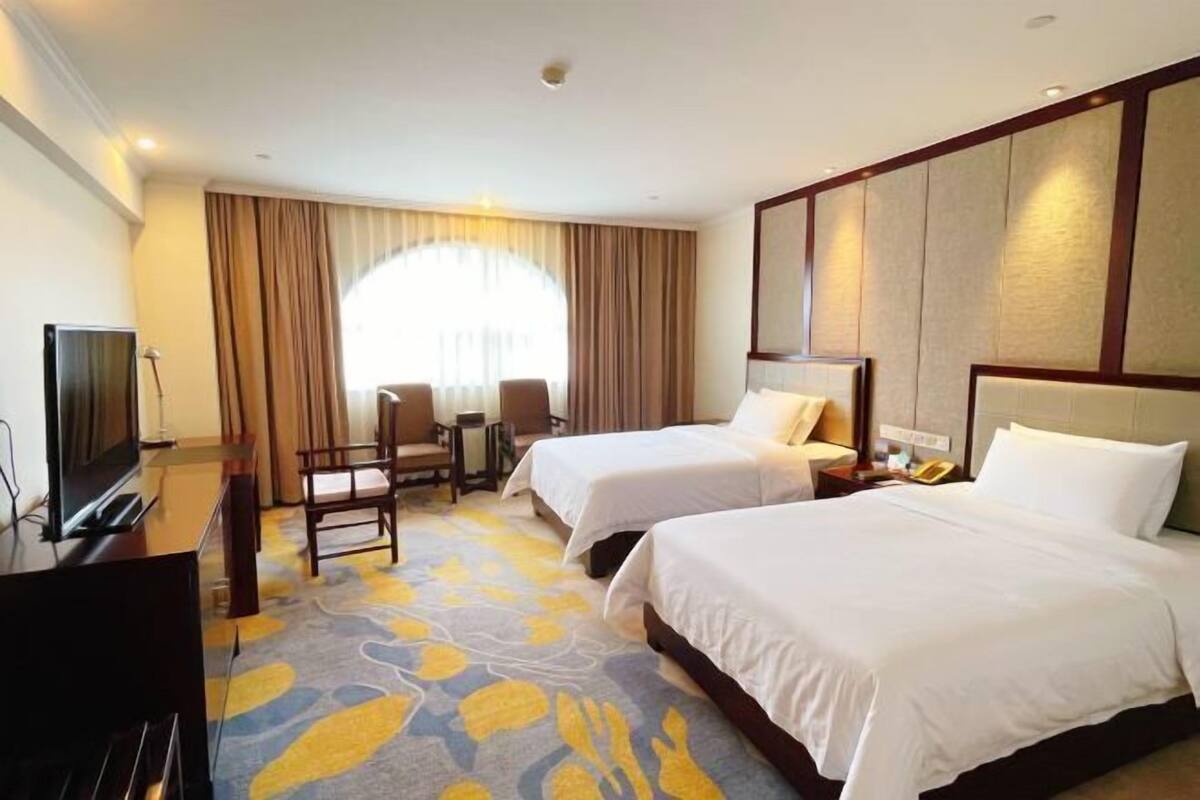 A hotel room with two beds, a desk with a TV, a chair, and a large window with curtains.