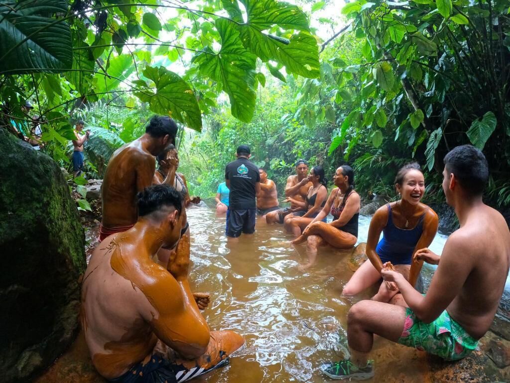 Un grupo de personas sentadas y de pie en un área natural y boscosa con un cuerpo de agua.