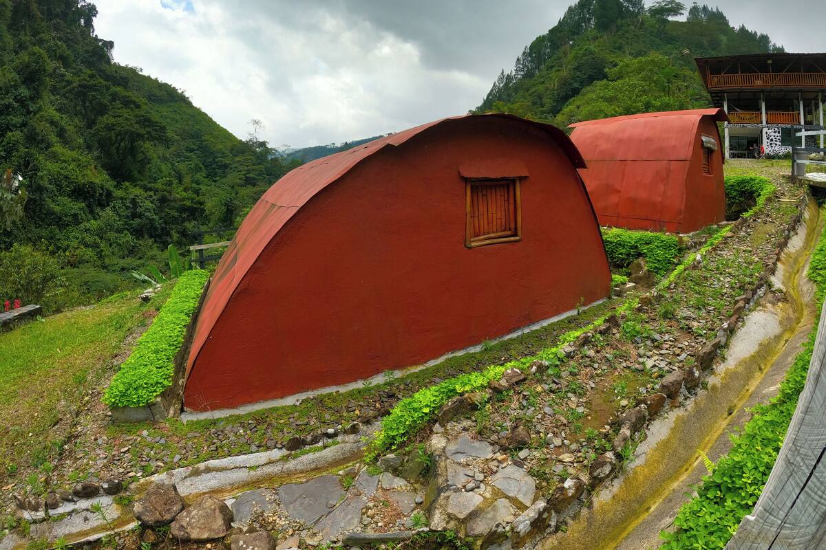 A red-roofed building with a small window, surrounded by greenery and a stone pathway.