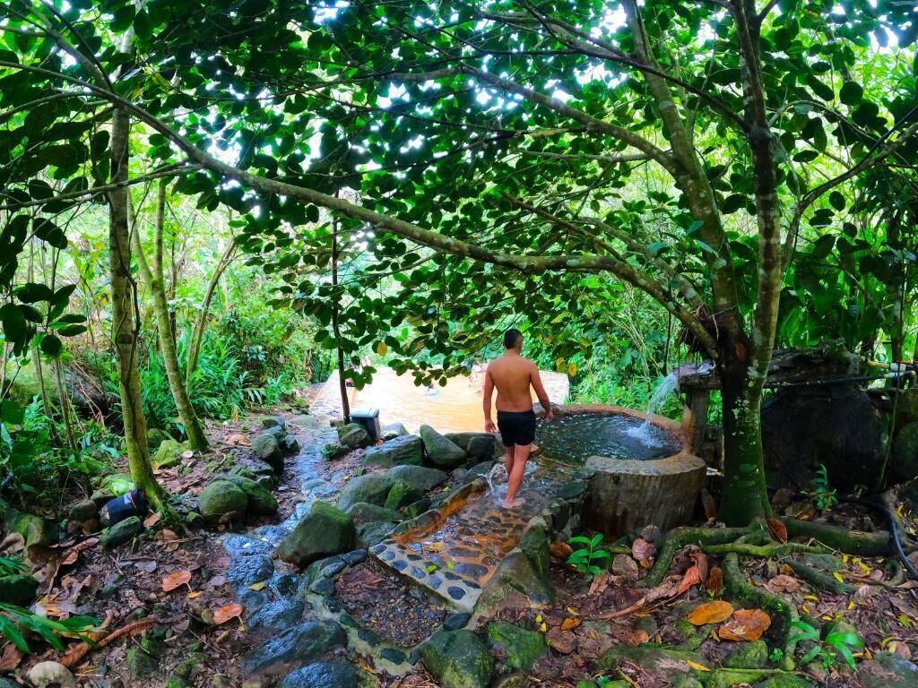 Una persona camina por un sendero de piedra hacia un manantial termal natural rodeado de un bosque denso.