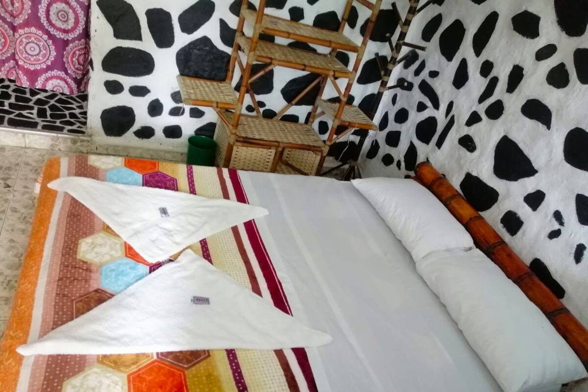 A bed with a white mattress and pillows, a colorful geometric-patterned rug, and a bamboo shelf unit against a wall with a black and white cow print design.