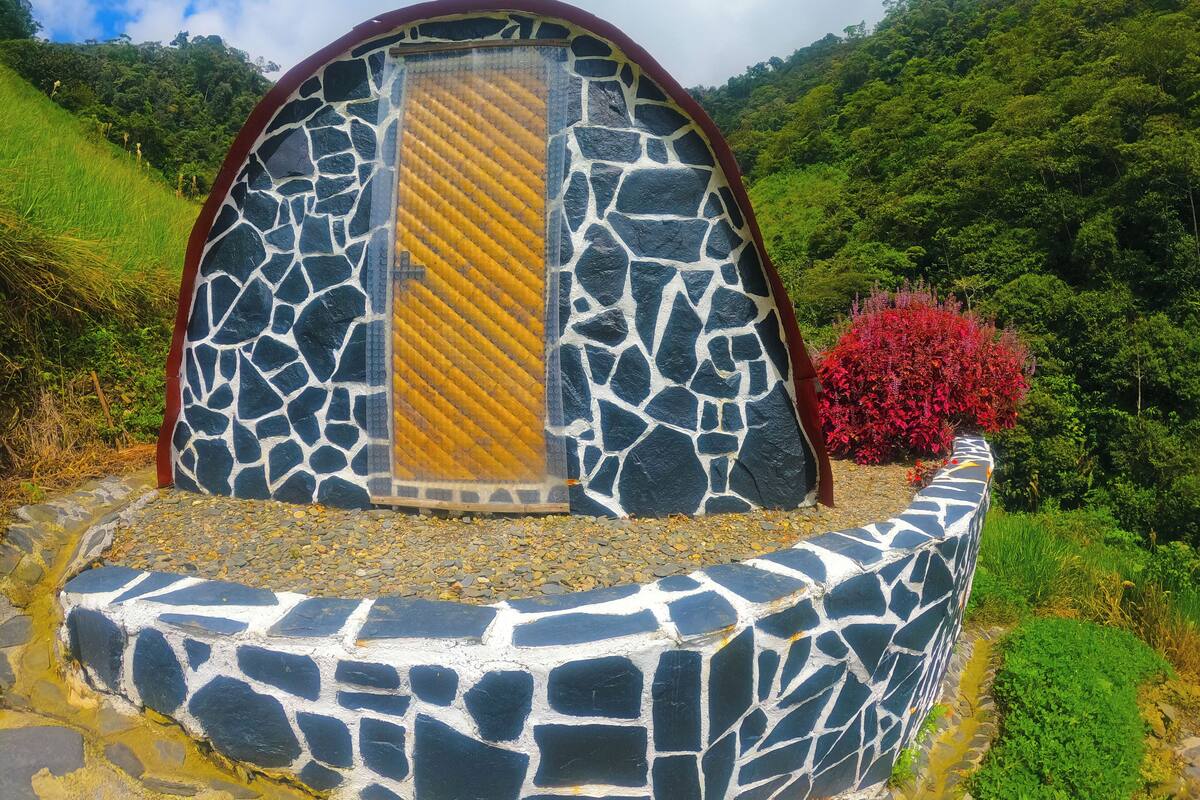 A stone structure with a curved roof and a central yellow and black striped panel, surrounded by greenery and a red bush.