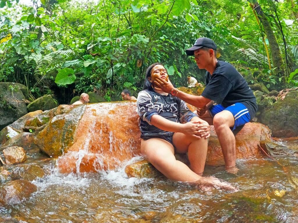 A person sitting on a rock by a flowing stream in a lush, green environment.