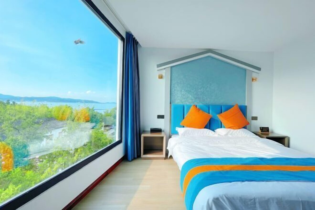 Habitación de hotel con un ventanal grande, una cama con ropa de cama azul y naranja, y una mesita de noche con lámpara.