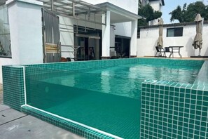 En rektangulär swimmingpool med en kant av kakel, omgiven av en vit byggnad med en täckt uteplats.